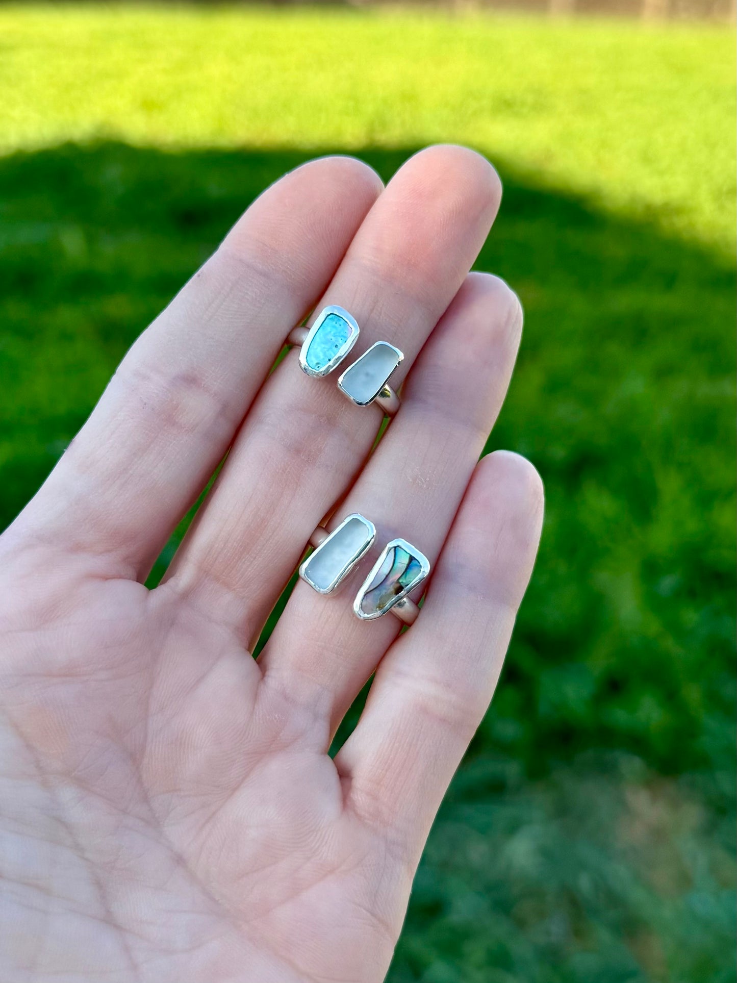 Two sea glass adn abalone shell silver bypass rings on a hand against a blurred green background