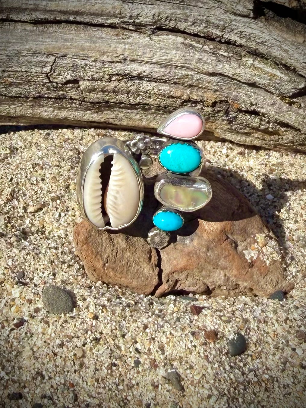 Ring with seashells and turquoise stones on a sandy surface with wood in the background