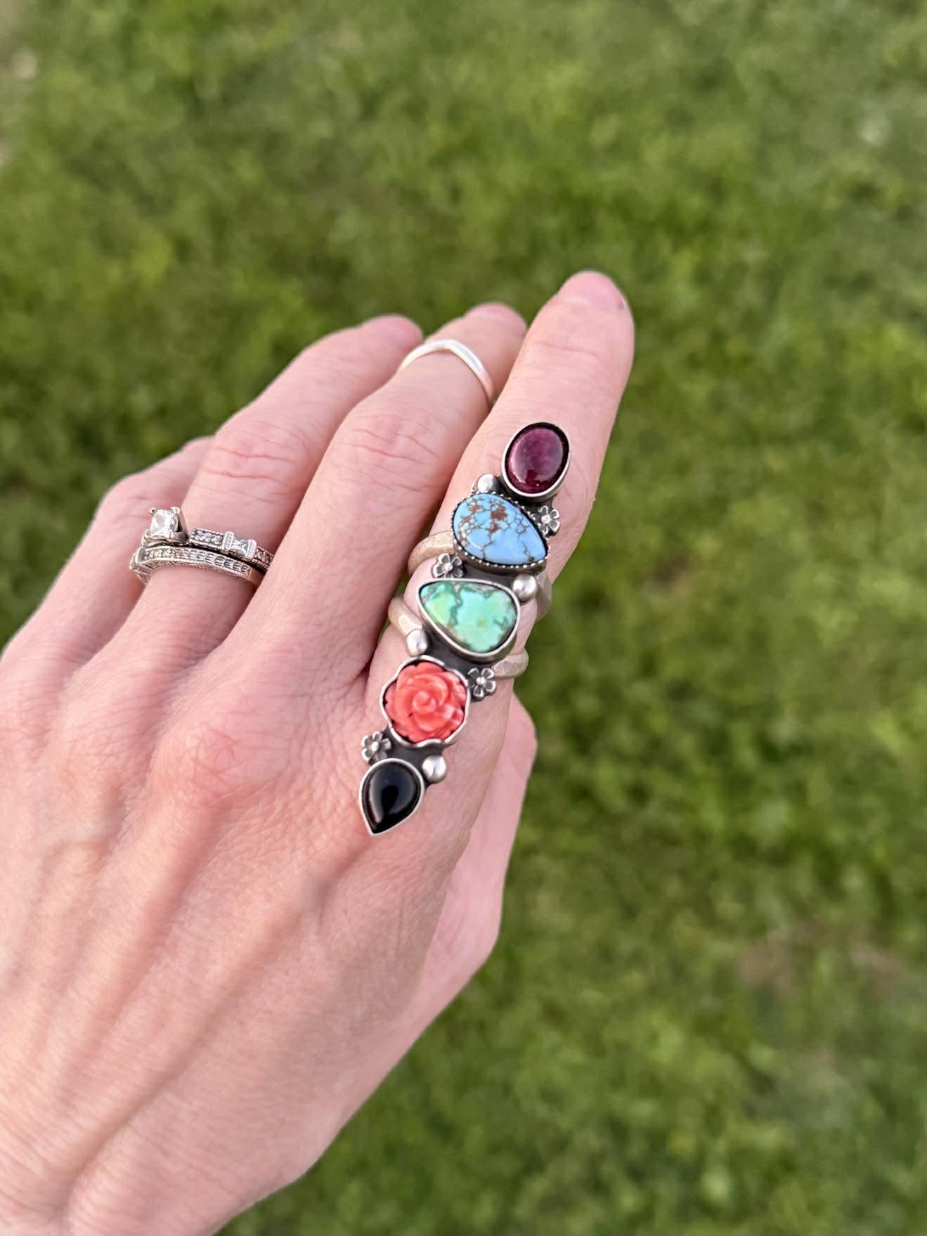 Hand wearing multiple rings with colorful gemstones against a grassy background