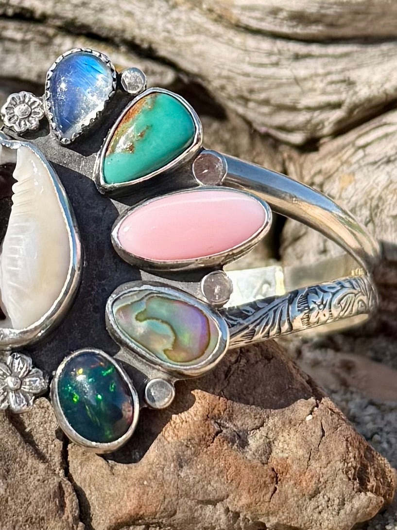close up of cowrie shell abalone pink conch opal turquoise and moonstone gemstone cuff bracelet on a piece of drift wood