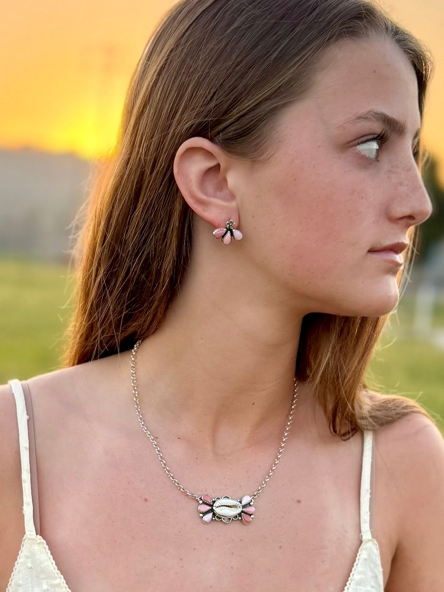Woman wearing a cowrie and ombre conch necklace and earrings to match outdoors with a sunset background