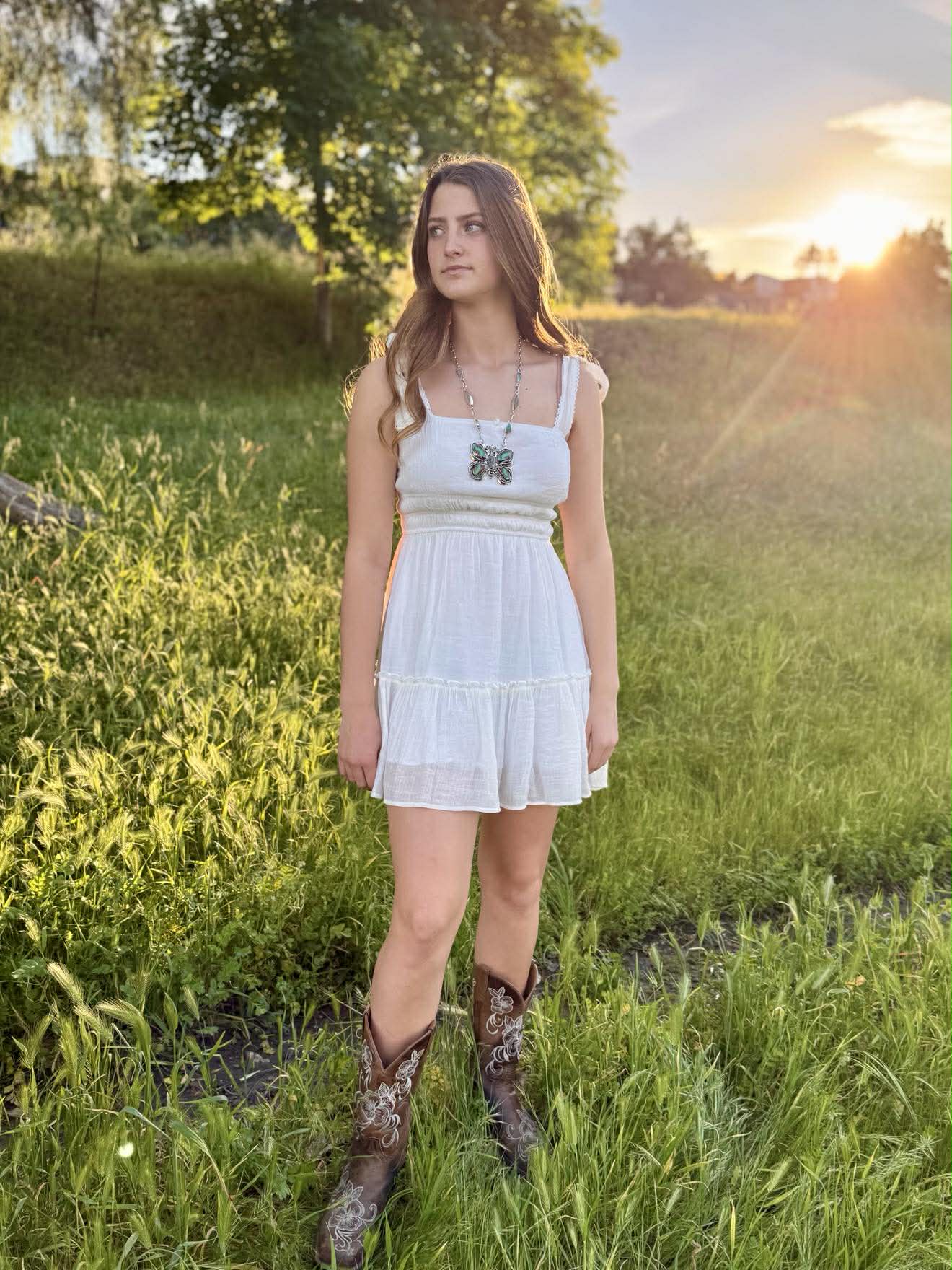Woman in a white dress and cowboy boots standing in a grassy field with sunset in the background
