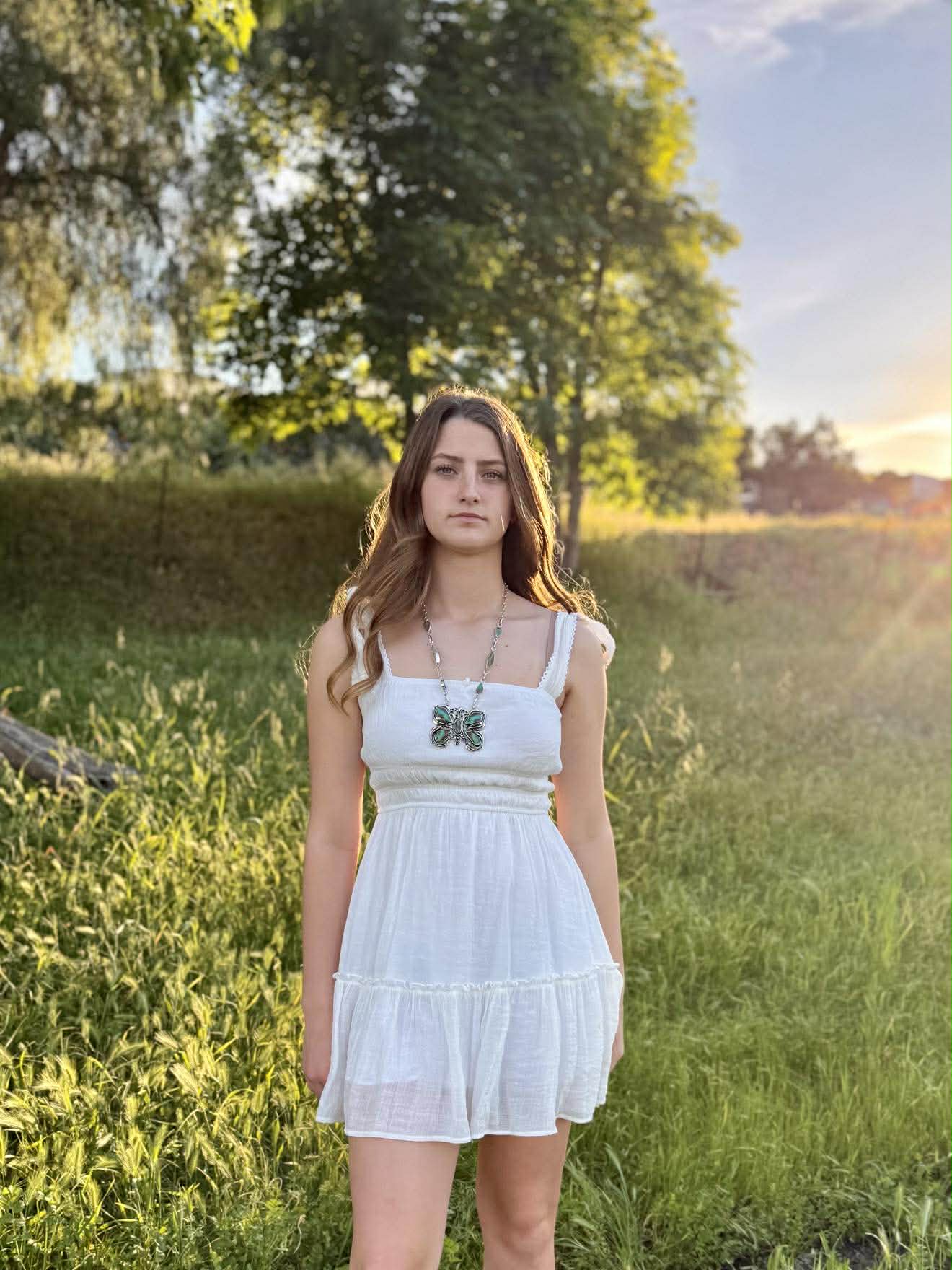 Woman in a white dress standing in a grassy field with trees and sunset in the background